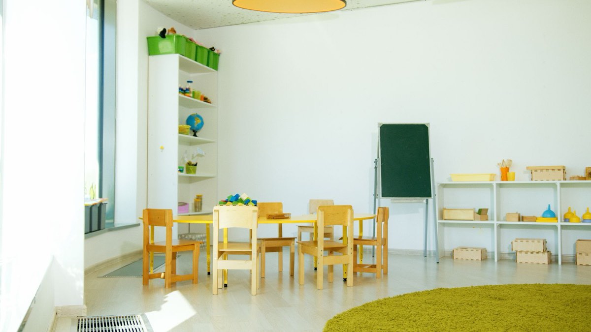 A sunlit nursery room with a low yellow table, small wooden chairs, a chalkboard easel and low shelves of wooden toys.
